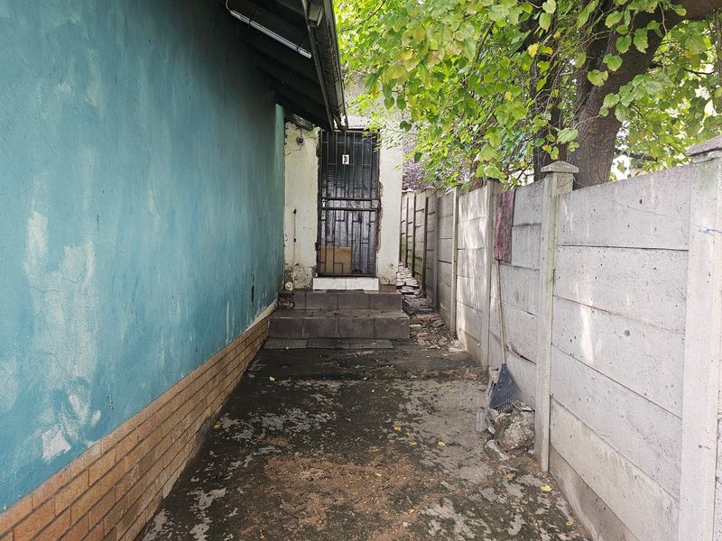 A narrow rear exterior walkway featuring a security gate, concrete boundary wall, and tiled steps leading to the entrance.