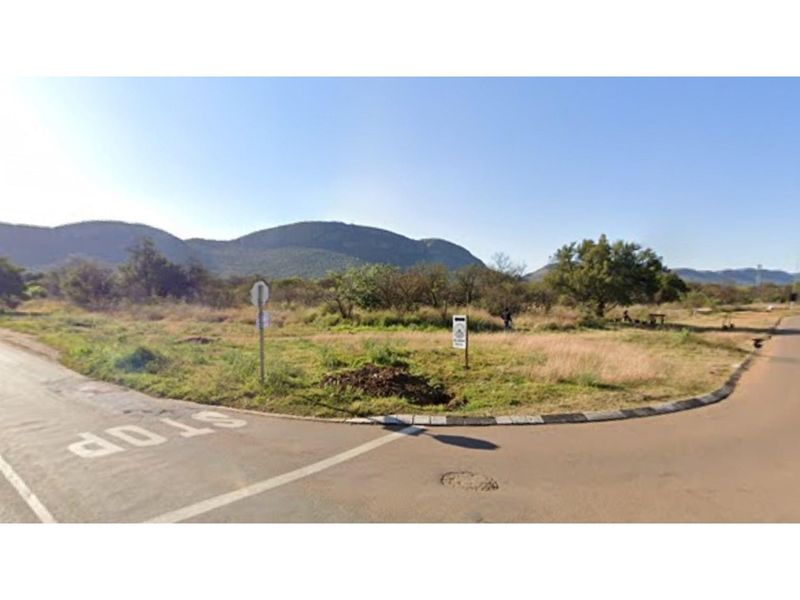 A street-level view of a vacant residential plot situated at a road intersection, featuring natural vegetation and mountain views in the background.