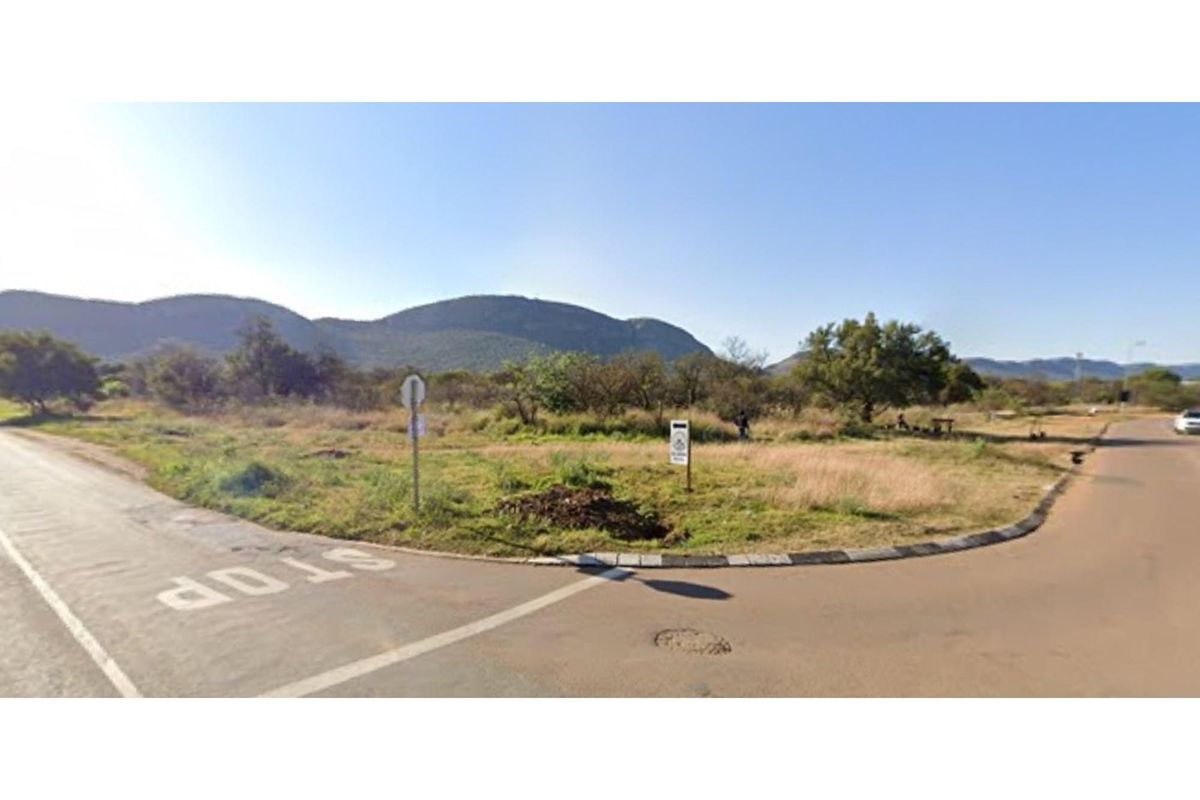 A street-level view of a vacant residential plot situated at a road intersection, featuring natural vegetation and mountain views in the background.