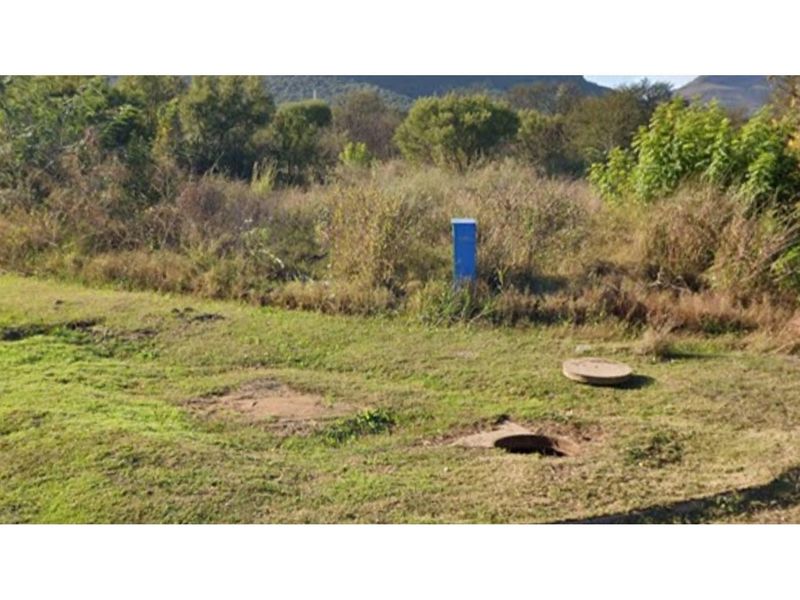 A view of the vacant residential land featuring natural vegetation, a utility connection box, and visible manhole covers on the site.