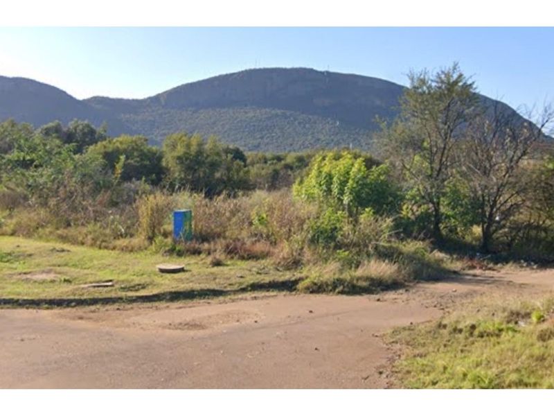 A view of a vacant residential plot featuring natural vegetation, a utility box, and a manhole cover with a mountain backdrop.