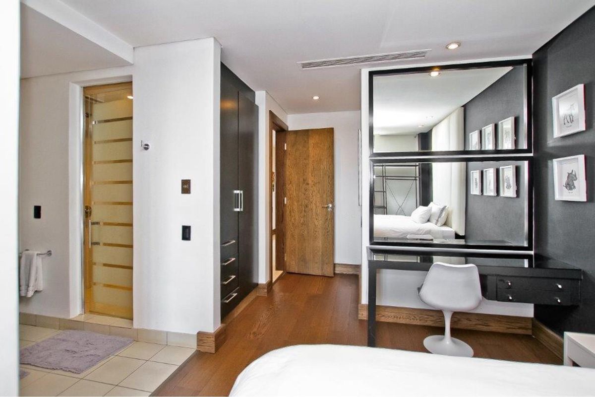 A modern bedroom featuring built-in dark cabinetry, wooden flooring, and an integrated vanity area with a large mirror, adjacent to a private bathroom.