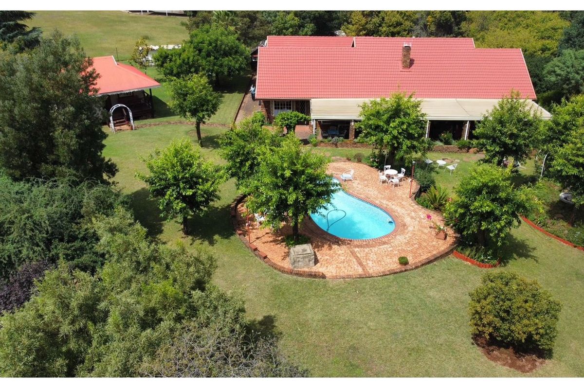Aerial view of a small holding featuring a main residence with a red roof, a swimming pool with brick paving, and expansive landscaped gardens.