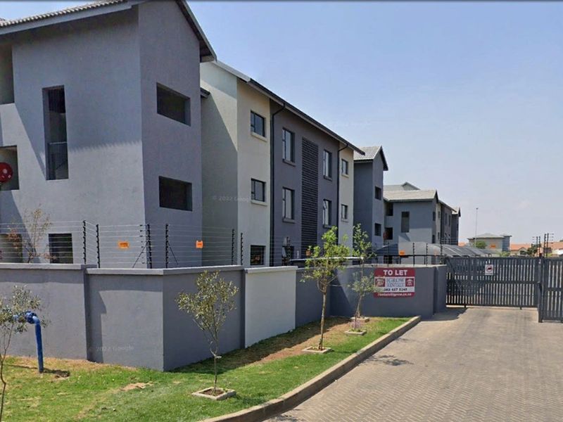 Exterior view of a modern multi-story apartment complex featuring paved driveway access, perimeter walling, and security electric fencing.