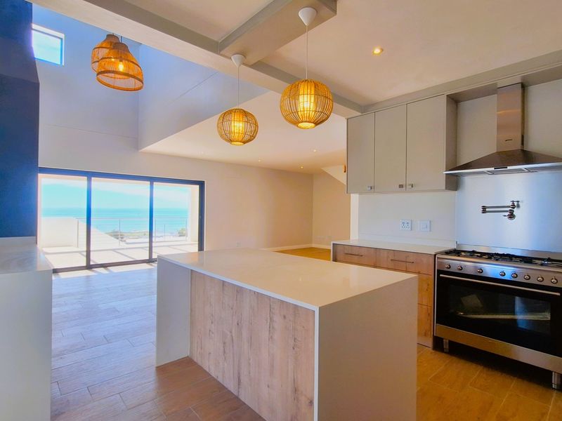 Modern open-plan kitchen and living area featuring a central island, high-end built-in appliances, and large sliding doors opening to a scenic coastal view.