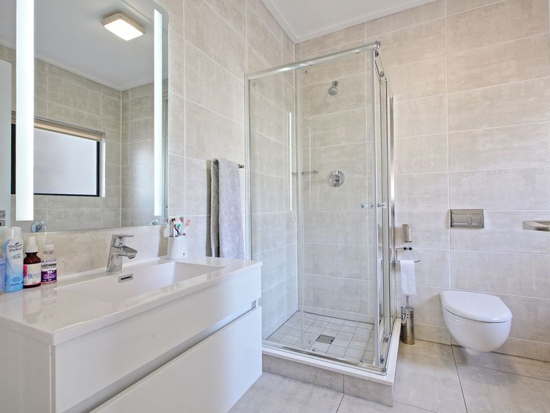 Modern bathroom featuring a wall-mounted toilet, glass shower enclosure, vanity with integrated basin, and a sleek backlit mirror with neutral floor-to-ceiling tiling.