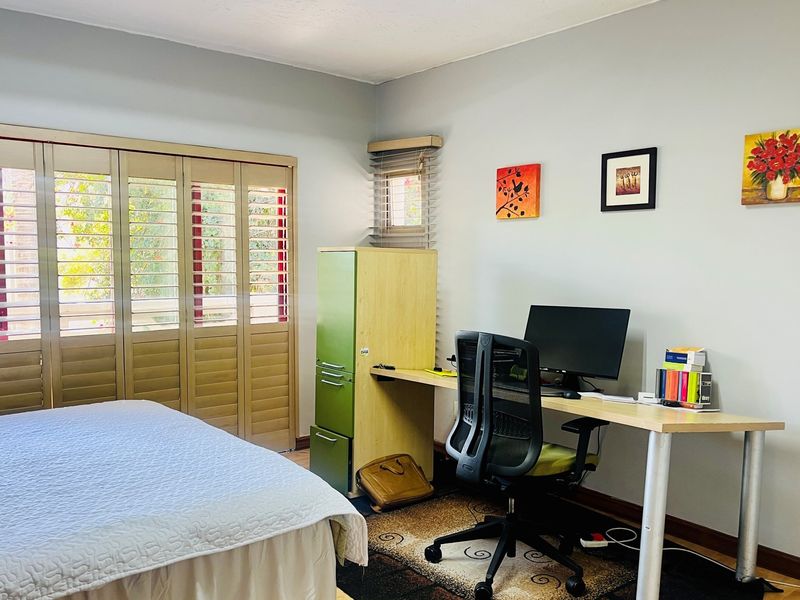 A functional bedroom featuring light-toned walls, wooden plantation shutters, and a built-in desk area, suitable for use as a home office or guest room.