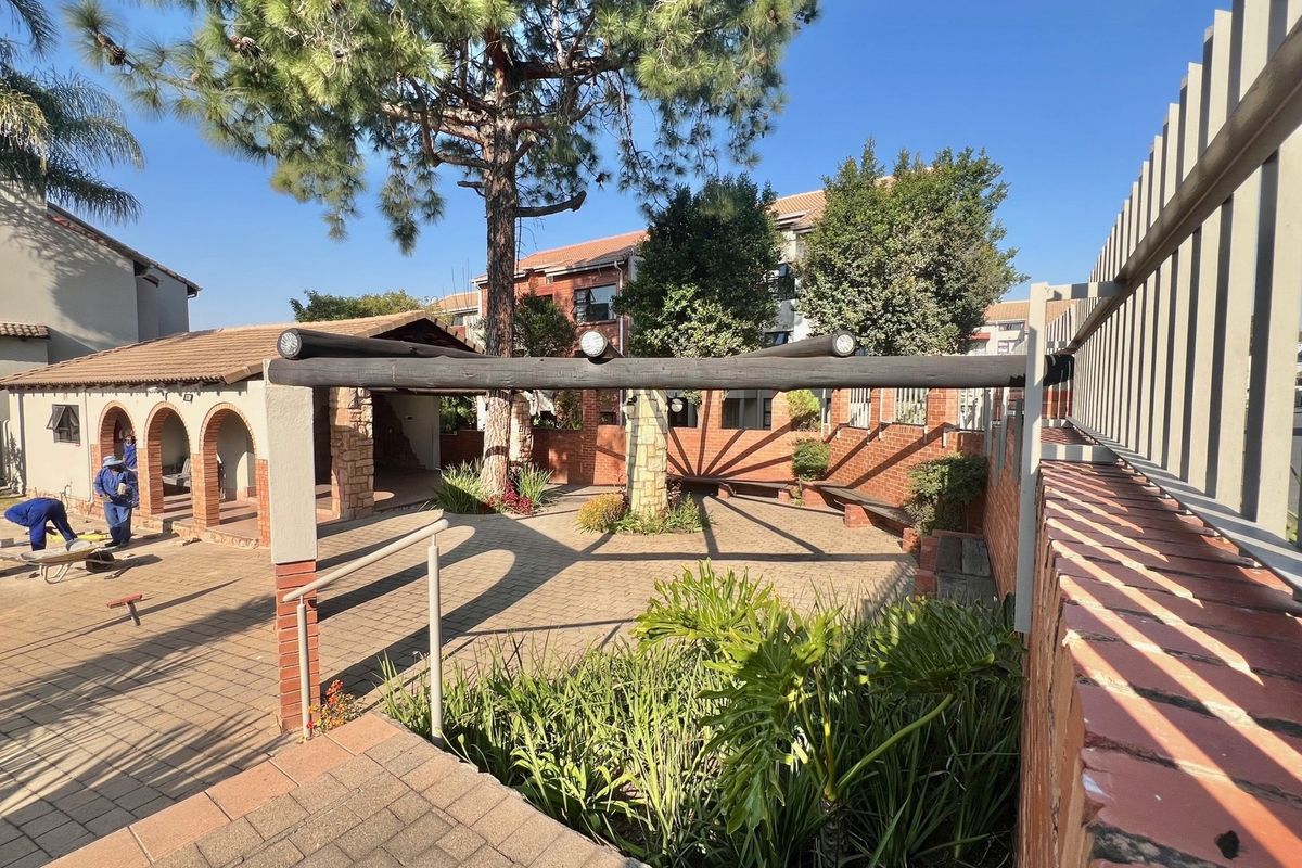 A communal outdoor courtyard area featuring paved flooring, a timber pergola structure, and established garden beds within an apartment complex setting.