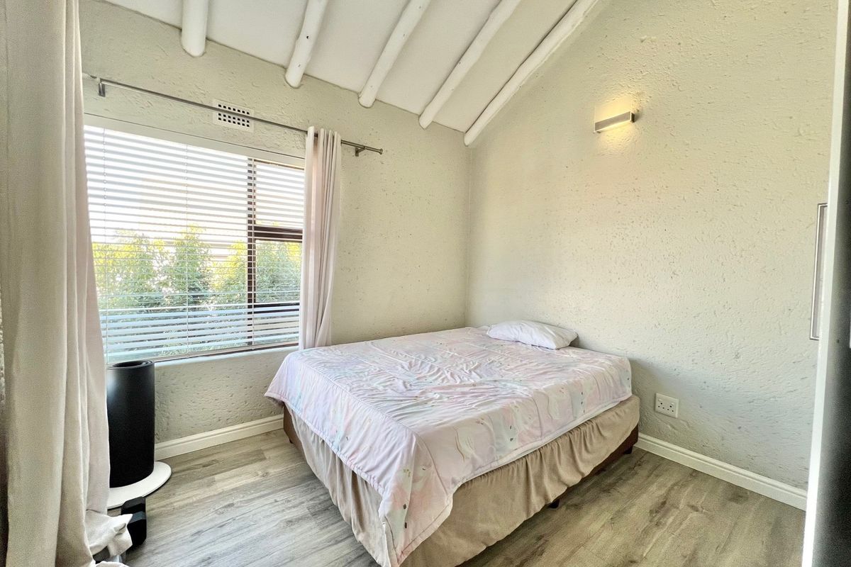 A bedroom featuring vaulted ceilings with exposed beams, wood-look flooring, and a large window with blinds.