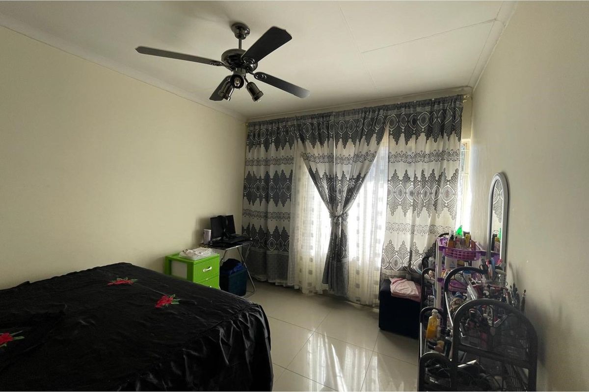 A standard bedroom featuring tiled flooring, a ceiling fan, and large windows covered by curtains.