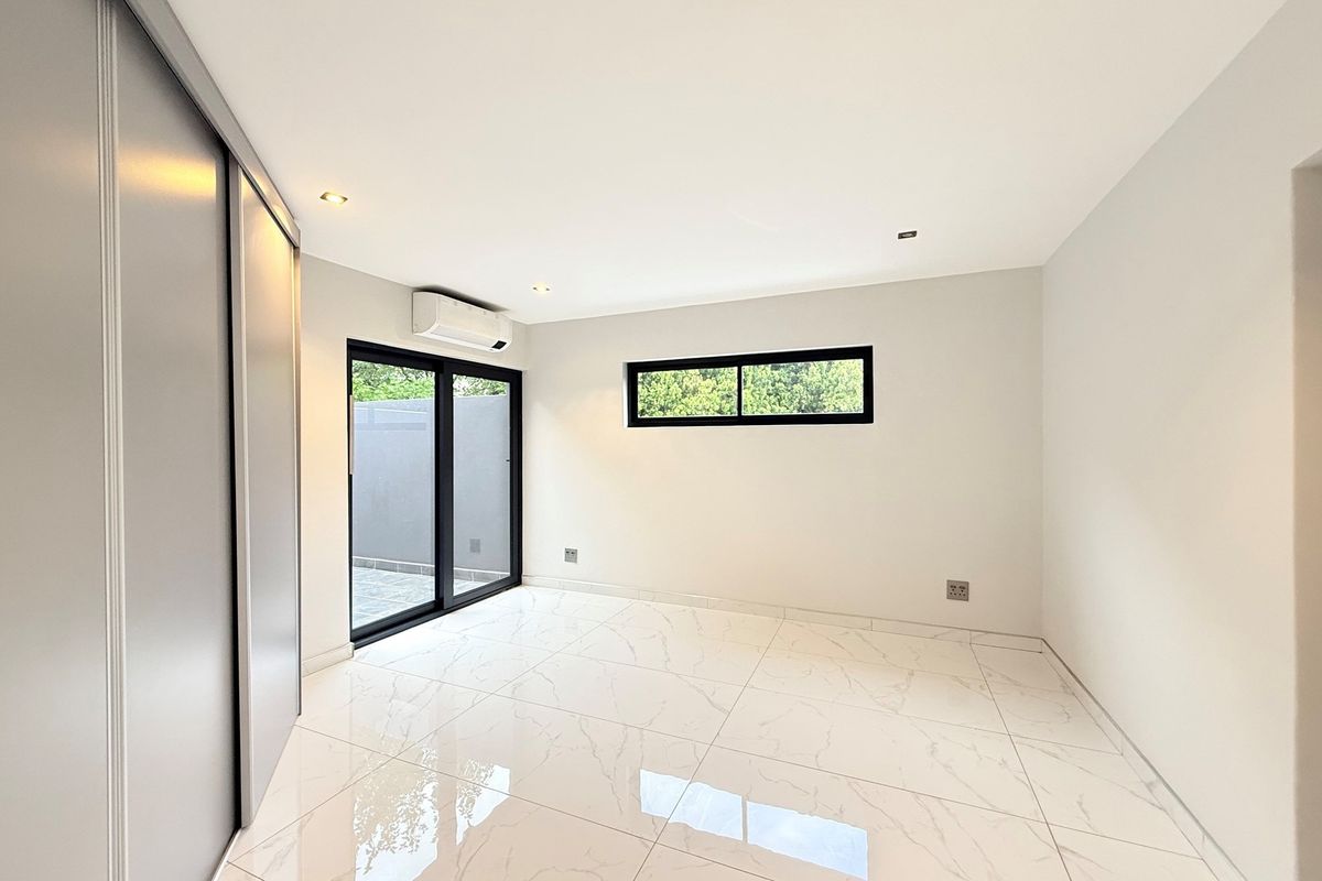 A bright, modern bedroom featuring polished marble-look floor tiles, built-in sliding door cupboards, a wall-mounted air conditioning unit, and black-framed windows and doors.