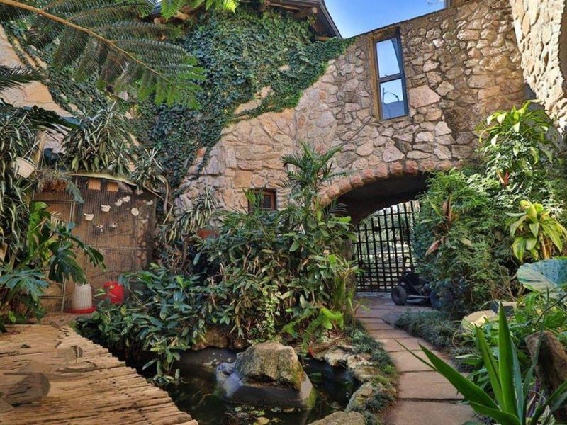 A charming stone-walled courtyard featuring a tranquil water feature, lush landscaping, and a rustic stone archway leading through to a gated entrance.