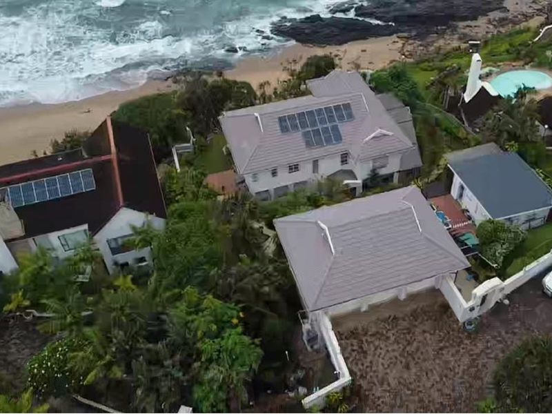 Aerial view of a coastal residential property featuring tiled roofs, solar panel installations, and proximity to the beach.