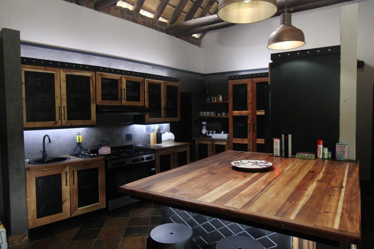 A rustic-industrial style kitchen featuring custom timber cabinetry, a large wooden island, exposed thatch roof beams, and integrated lighting.