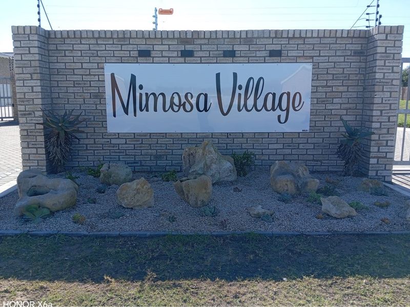 The entrance to Mimosa Village features a brick boundary wall with electric fencing, a secure metal gate, and a landscaped rock garden.