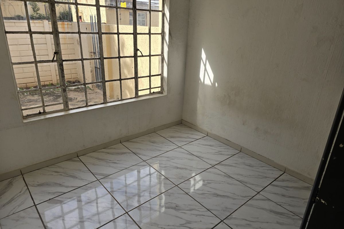 An empty bedroom featuring white tiled flooring and a large steel-framed window, requiring cosmetic updates to the walls and window frame.