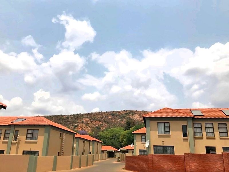 A view of a residential complex featuring multi-story units with terracotta tiled roofs, solar panels, and paved access roads against a mountain backdrop.
