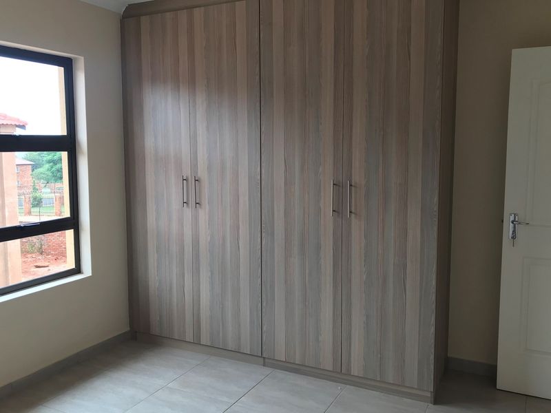 A clean, empty bedroom featuring modern wood-finish built-in cupboards, neutral tiled flooring, and a large window providing natural light.