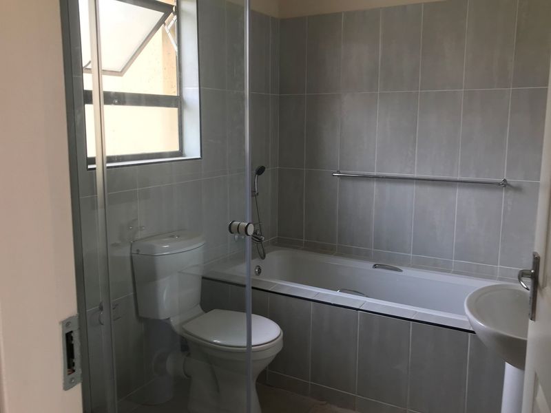 A clean, modern bathroom featuring a bathtub, pedestal basin, toilet, and neutral wall tiling with a glass shower screen.