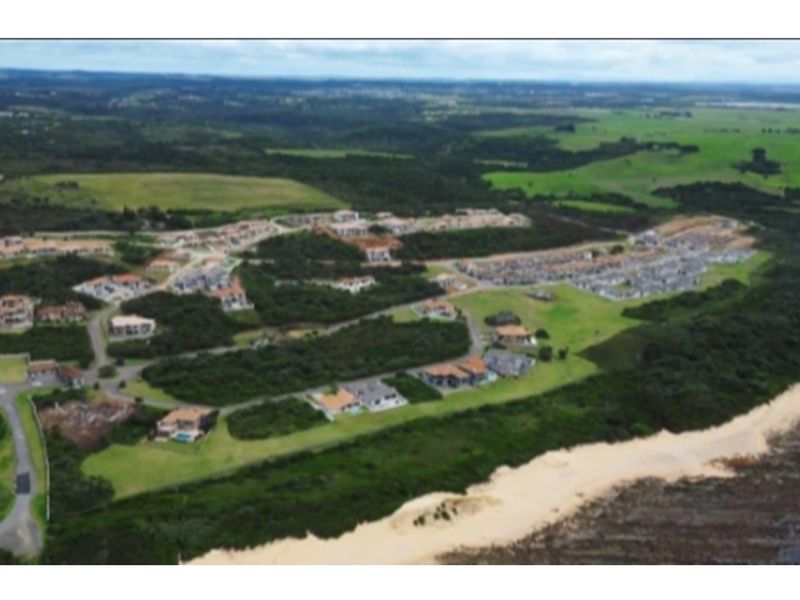 An aerial view of a coastal residential estate, showcasing the surrounding landscape, road infrastructure, and proximity to the beach.