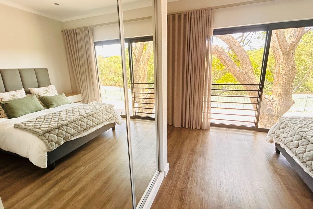 A bright bedroom featuring wood-look flooring, built-in mirrored sliding wardrobes, and large sliding glass doors that open to a balcony with tree views.