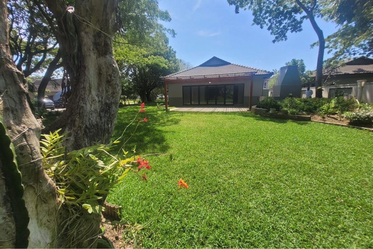 A spacious rear garden featuring a well-maintained lawn, mature trees, and a covered patio area with a built-in outdoor fireplace.