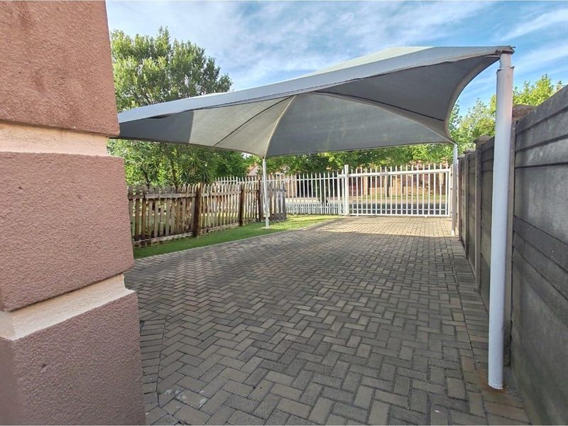 A paved driveway area featuring a shade sail structure, enclosed by a combination of timber and concrete boundary fencing with a metal gate.