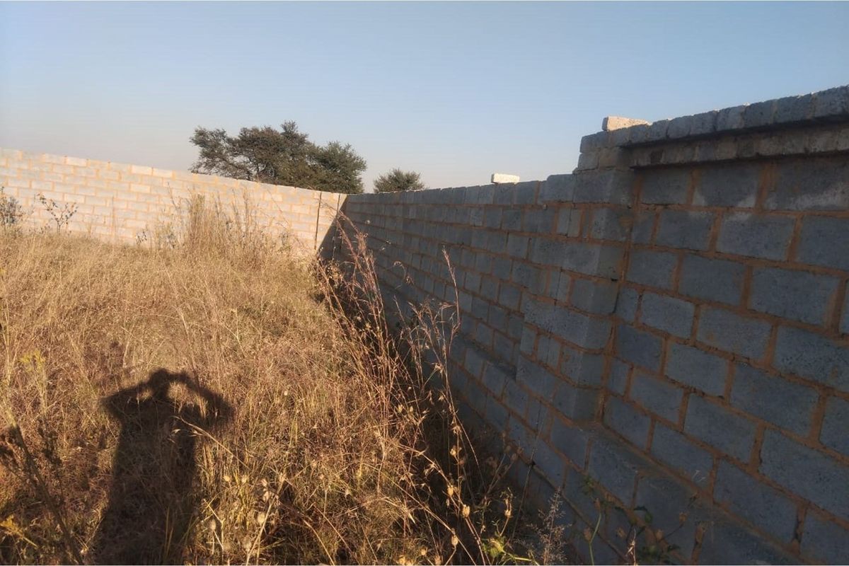 A view of a vacant residential plot featuring a perimeter concrete block boundary wall and overgrown natural vegetation.