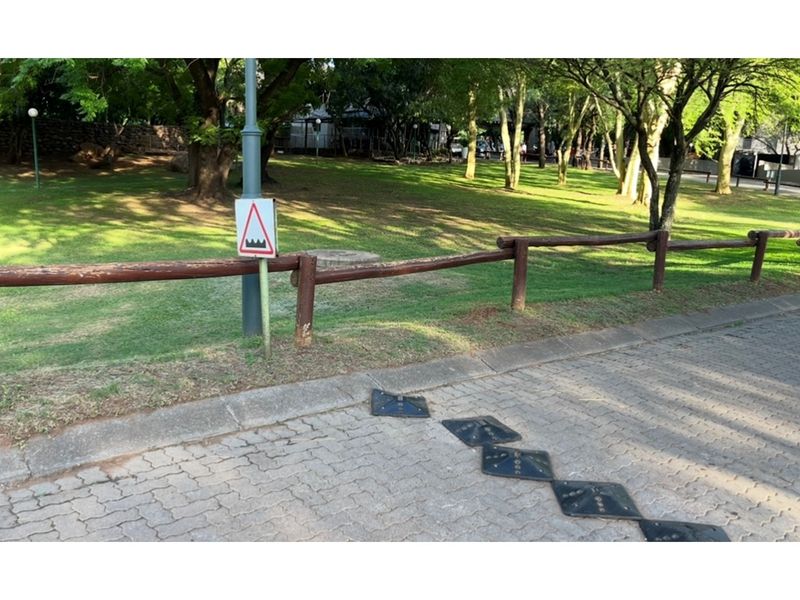 Street view of a residential area featuring a paved road with speed bumps, a timber fence, and a warning sign adjacent to a grassy verge.