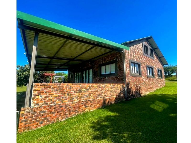 A rear view of the house featuring a face brick exterior, a covered patio area, and a corrugated metal roof set within a grassy garden.