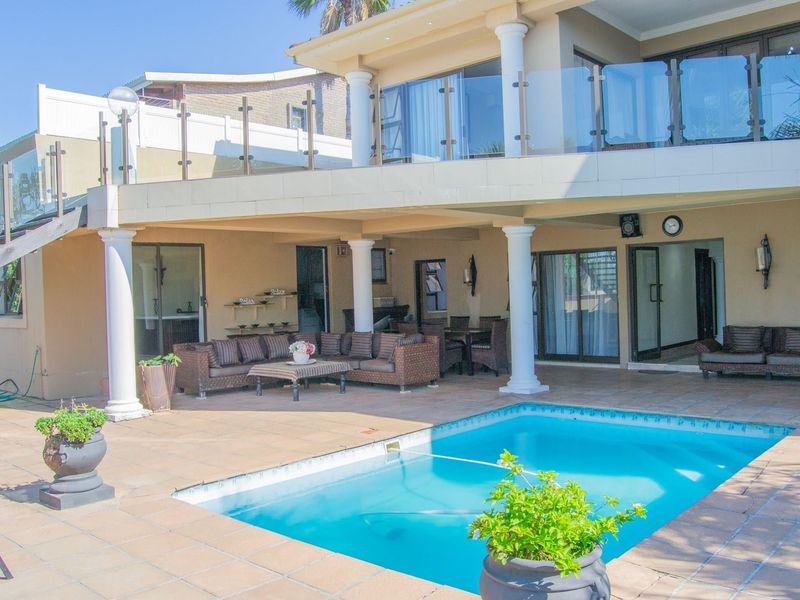 Spacious covered patio area overlooking a swimming pool, featuring tiled flooring, structural columns, and a glass balustrade on the upper level.