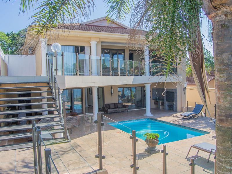 Rear exterior view featuring a swimming pool, tiled patio area, built-in braai, and a double-story facade with glass balustrades and an external staircase.