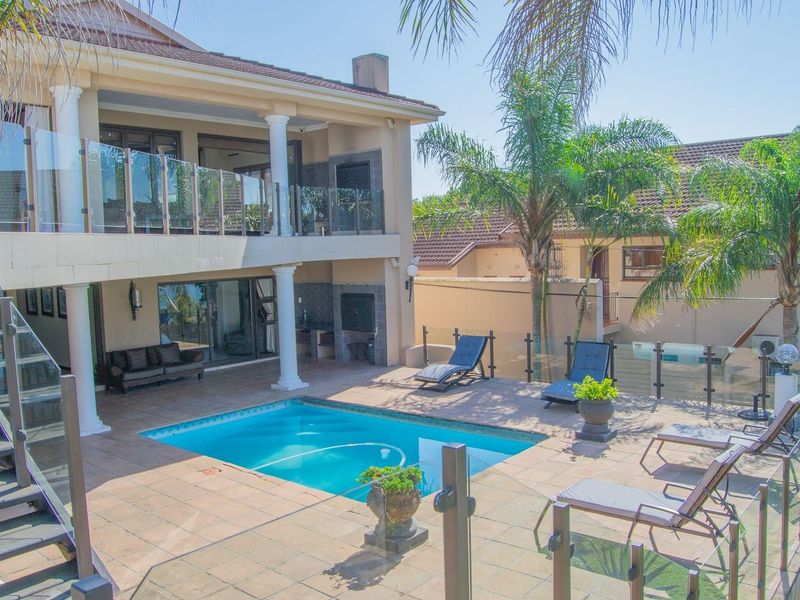 A spacious outdoor entertainment area featuring a swimming pool, tiled patio, covered terrace with built-in braai, and modern glass balustrades.