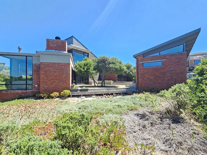 Rear view of a modern architectural home featuring exposed brickwork, large glass windows, and a timber deck set within a landscaped garden.