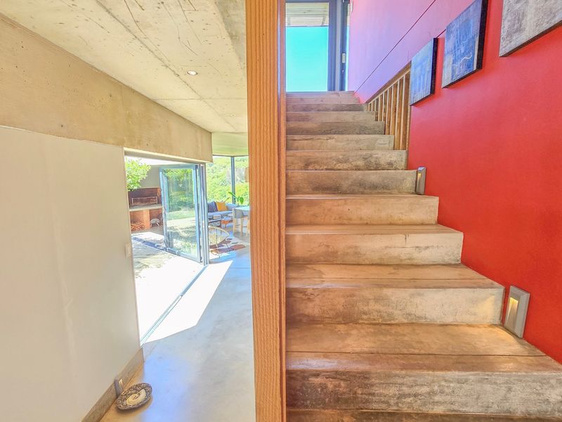 A modern hallway featuring a striking concrete staircase with integrated lighting, exposed concrete ceilings, and seamless access to an outdoor patio area.