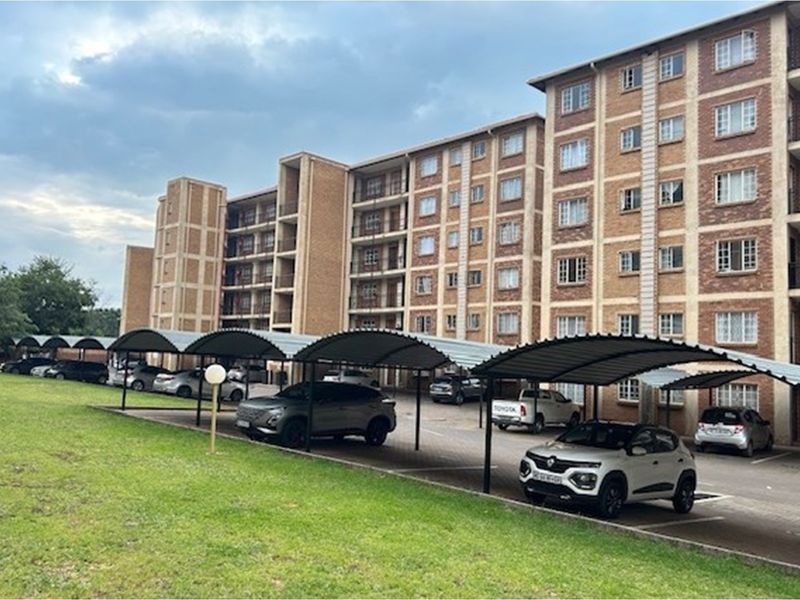 Exterior view of a multi-story brick apartment complex featuring covered parking bays and a paved driveway area.