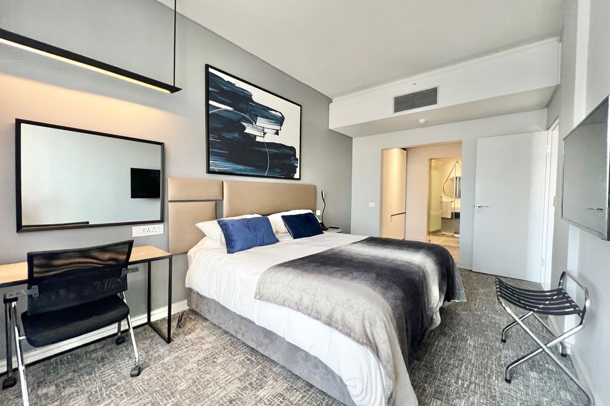 A modern bedroom featuring a built-in headboard, integrated desk area, and direct access to an en-suite bathroom with neutral finishes.