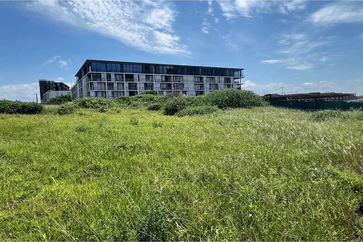 A view of a vacant residential plot featuring natural grass coverage with neighboring multi-story apartment buildings in the background.