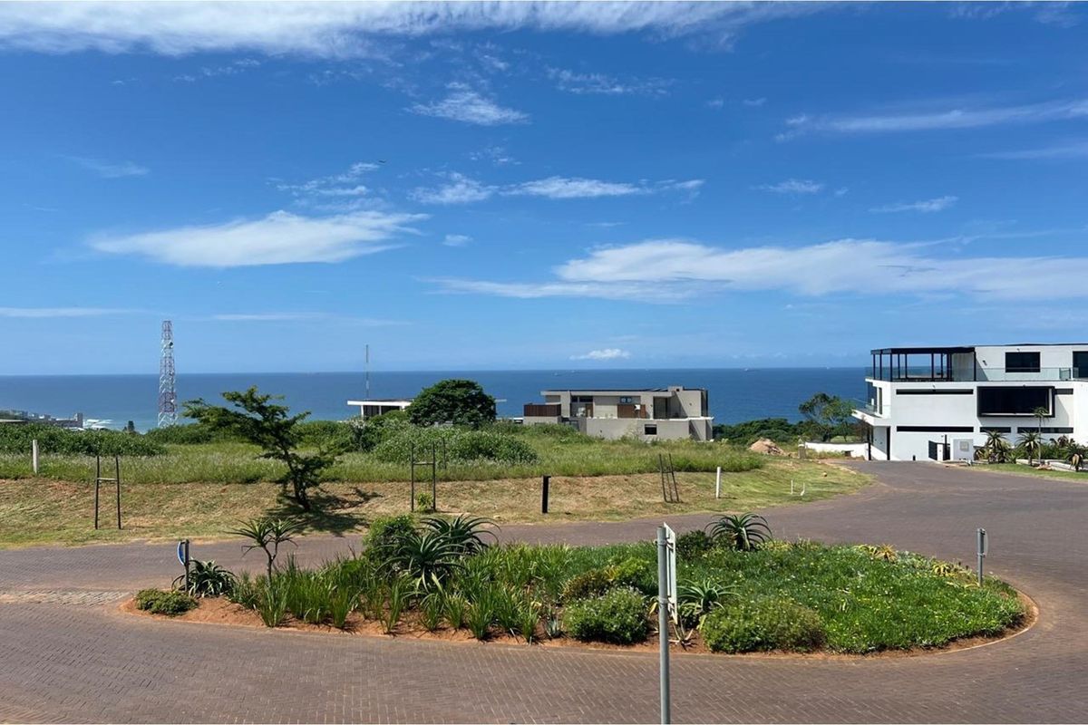 A view of a vacant residential plot in a coastal estate, featuring paved access roads, landscaped verge, and distant ocean views.