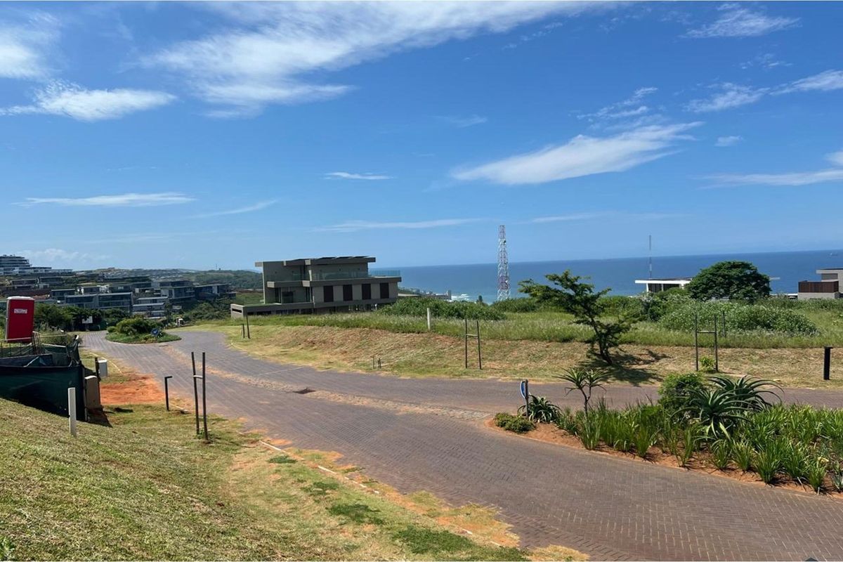 A view of a vacant residential plot featuring paved road access, established landscaping, and scenic coastal views in a developed estate setting.