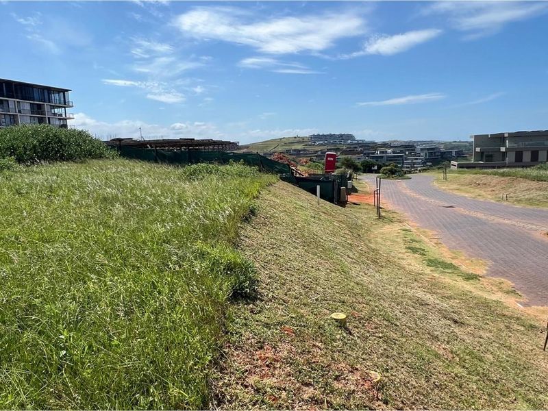 A view of a vacant residential plot featuring sloped terrain and adjacent paved road access within a developed estate.