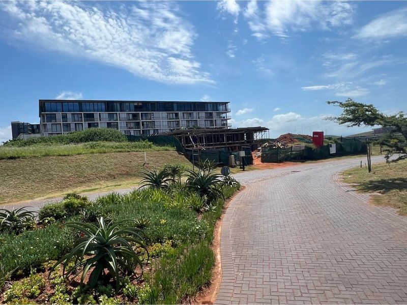 A view of a paved residential street leading towards a construction site and existing apartment complex in a coastal estate setting.