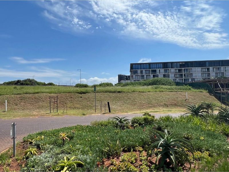 A view of a vacant residential plot featuring a paved driveway, landscaped verge, and a grassed embankment with surrounding estate development.