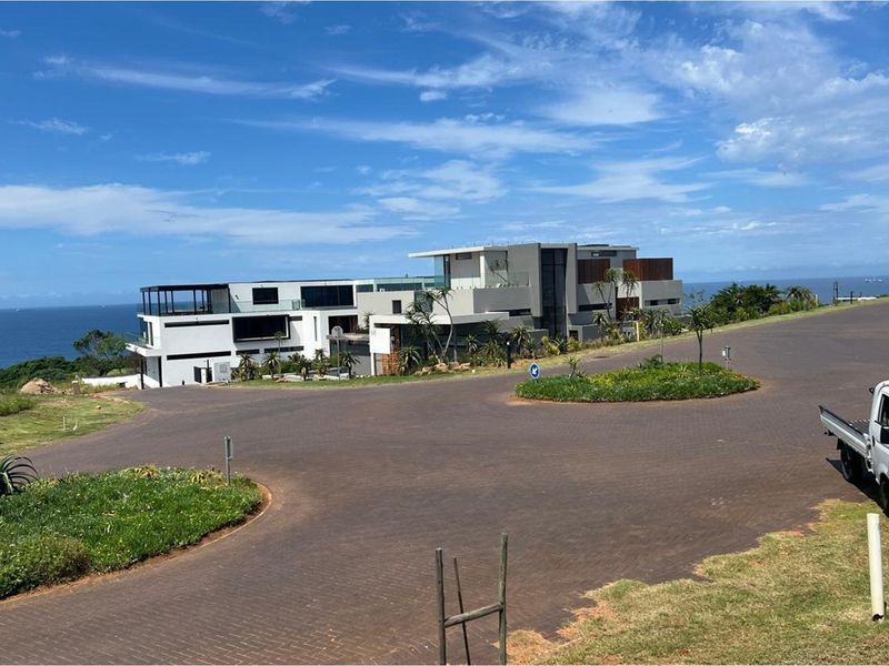 A street-level view of a modern, multi-level residential property situated in a coastal estate with paved roads and landscaped surroundings.