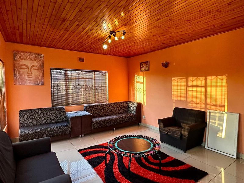 A living room featuring a warm timber tongue and groove ceiling, tiled flooring, and large windows with fitted venetian blinds.