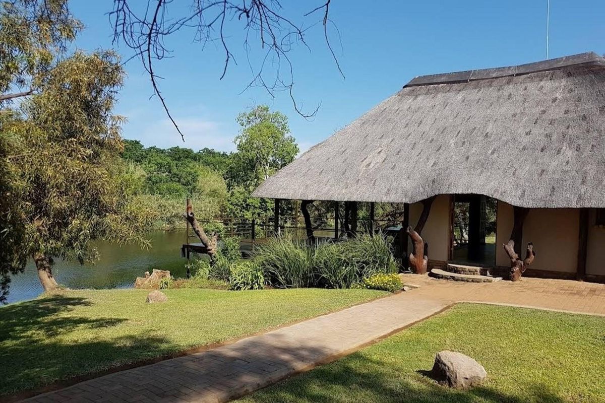 A scenic view of a thatched-roof dwelling situated by a tranquil lake, featuring a paved walkway and established garden surroundings.