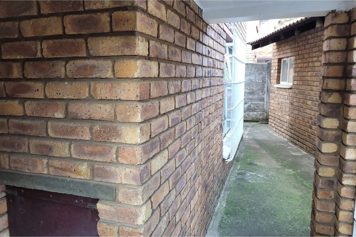 A narrow exterior side passage featuring face brick walls, a security gate, and a concrete pathway showing signs of weathering and moss.