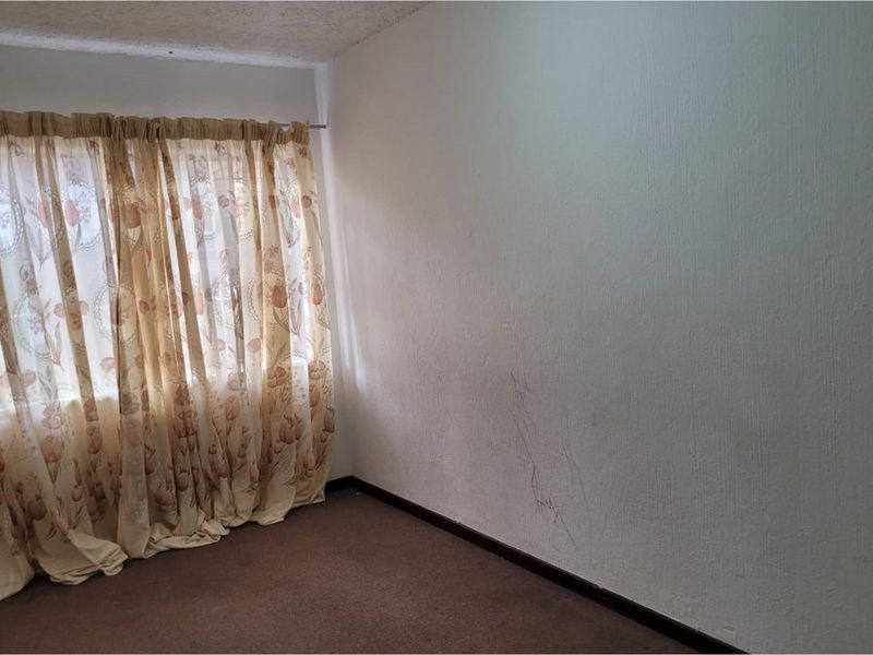 An empty bedroom featuring carpet flooring and a window with a curtain rod, showing signs of wear on the walls and floor.