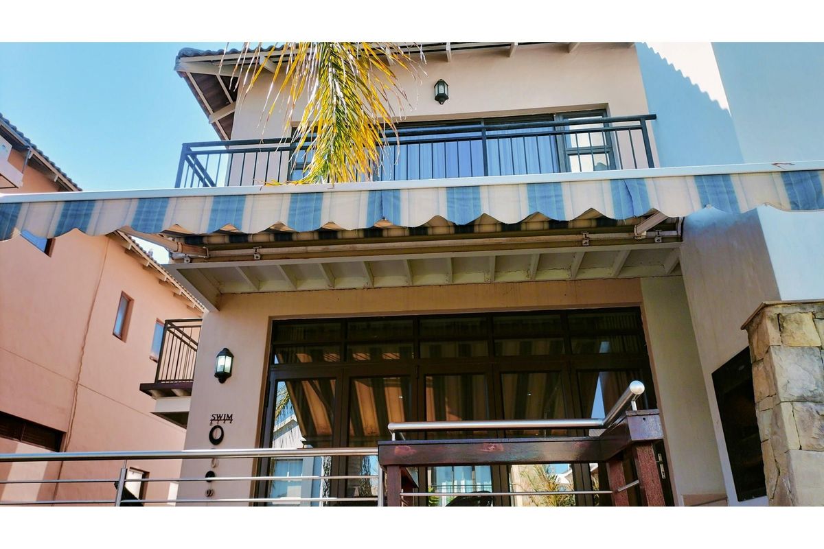 Rear exterior view featuring a covered patio with a retractable striped awning, a balcony above, and large glass sliding doors.