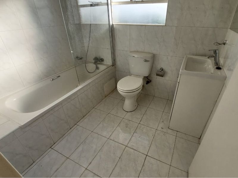 A functional bathroom featuring a bathtub with a glass screen, a toilet, and a vanity basin, finished with neutral wall and floor tiling.